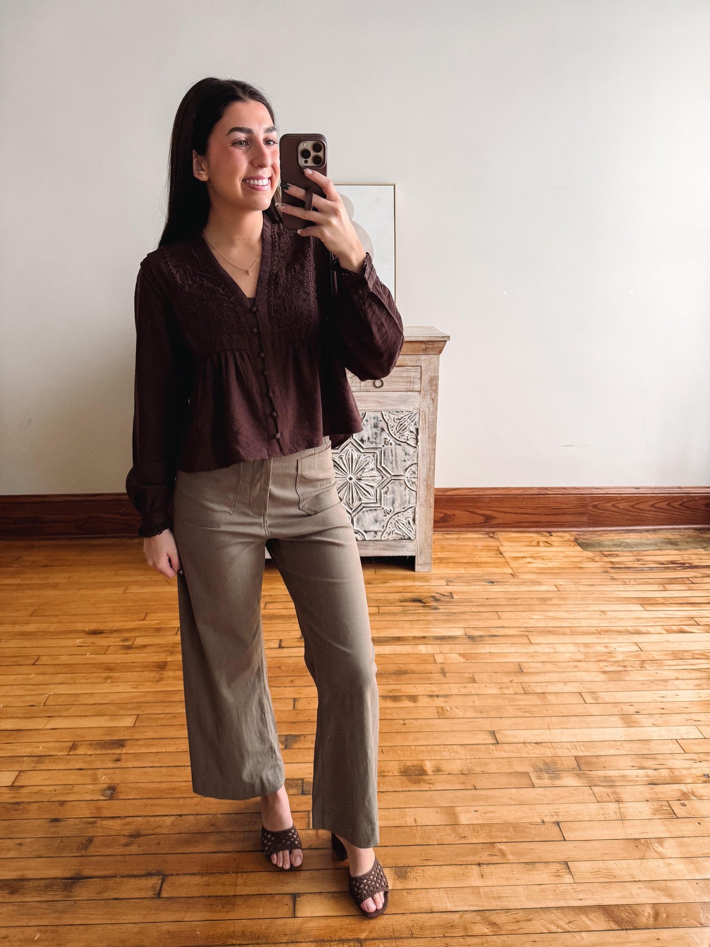 A woman takes a mirror selfie in a room with hardwood floors and a white wall, wearing a dark brown lace-trimmed long-sleeve blouse and olive green wide-leg pants.