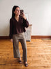 A woman takes a mirror selfie in a room with hardwood floors and a white wall, wearing a dark brown lace-trimmed long-sleeve blouse and olive green wide-leg pants.