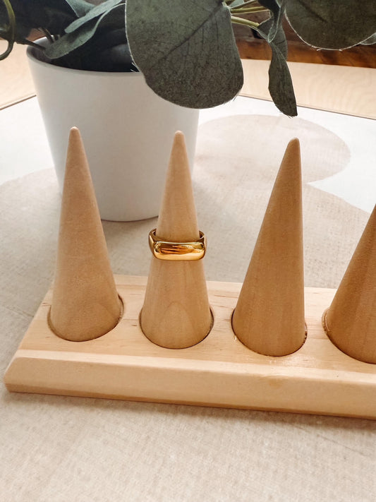 A gold ring displayed on one of four cone-shaped wooden ring stands set on a light wood base, with a white planter in the background.