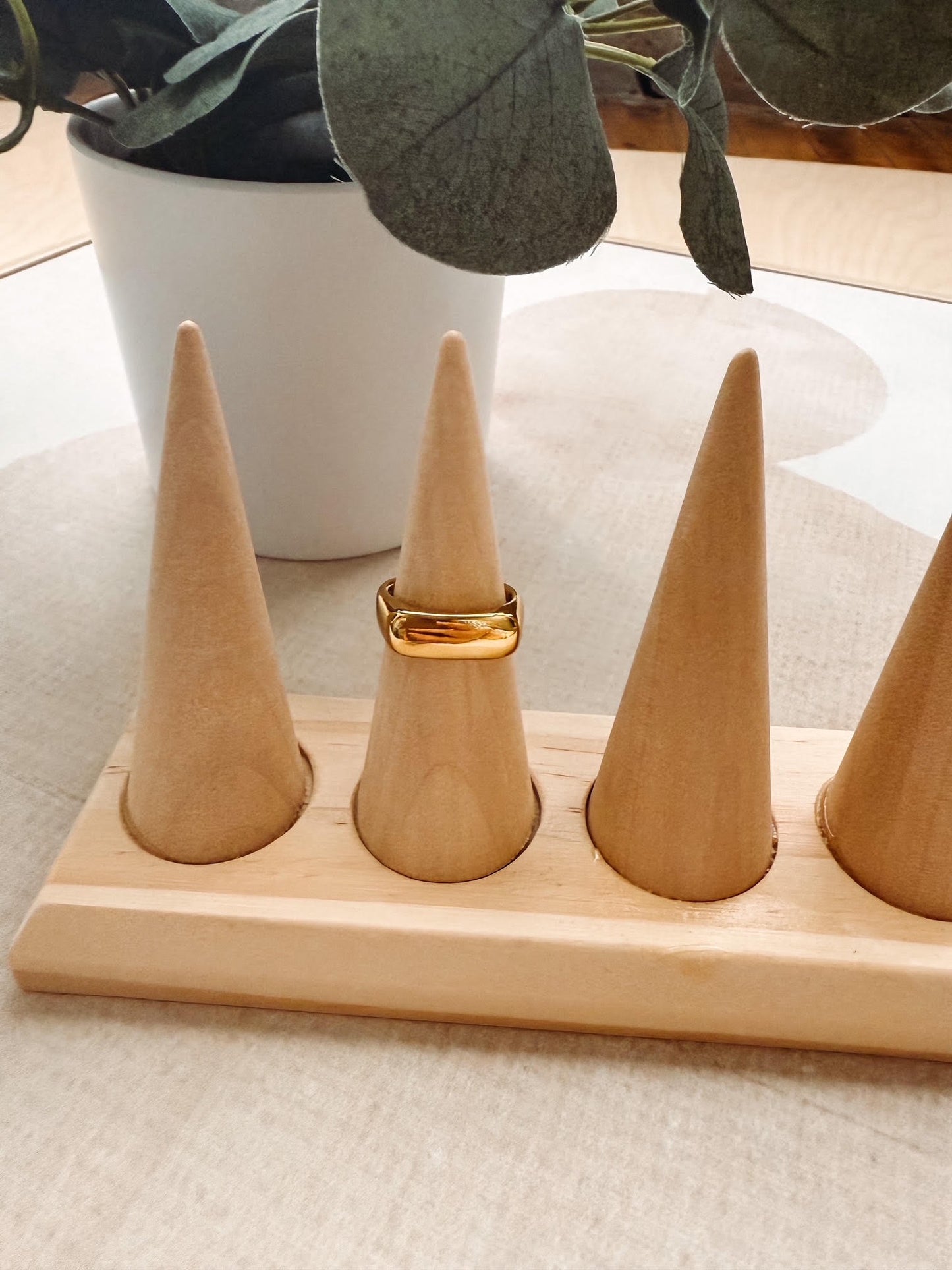 A gold ring displayed on one of four cone-shaped wooden ring stands set on a light wood base, with a white planter in the background.