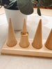 A gold ring displayed on one of four cone-shaped wooden ring stands set on a light wood base, with a white planter in the background.