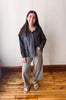 A woman modeling a dark charcoal gray button-down shirt and light beige wide-leg pants, standing on a wooden floor against a plain off-white wall.