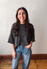 A woman with long dark hair stands indoors wearing a dark charcoal gray short-sleeve knit top and light blue jeans, with her hand in her pocket, against a plain off-white wall and wood floor.