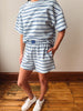A person wearing a matching blue and white striped lounge set, featuring an oversized short-sleeve t-shirt and coordinating shorts, paired with white sneakers, standing on a wooden floor against a beige wall.
