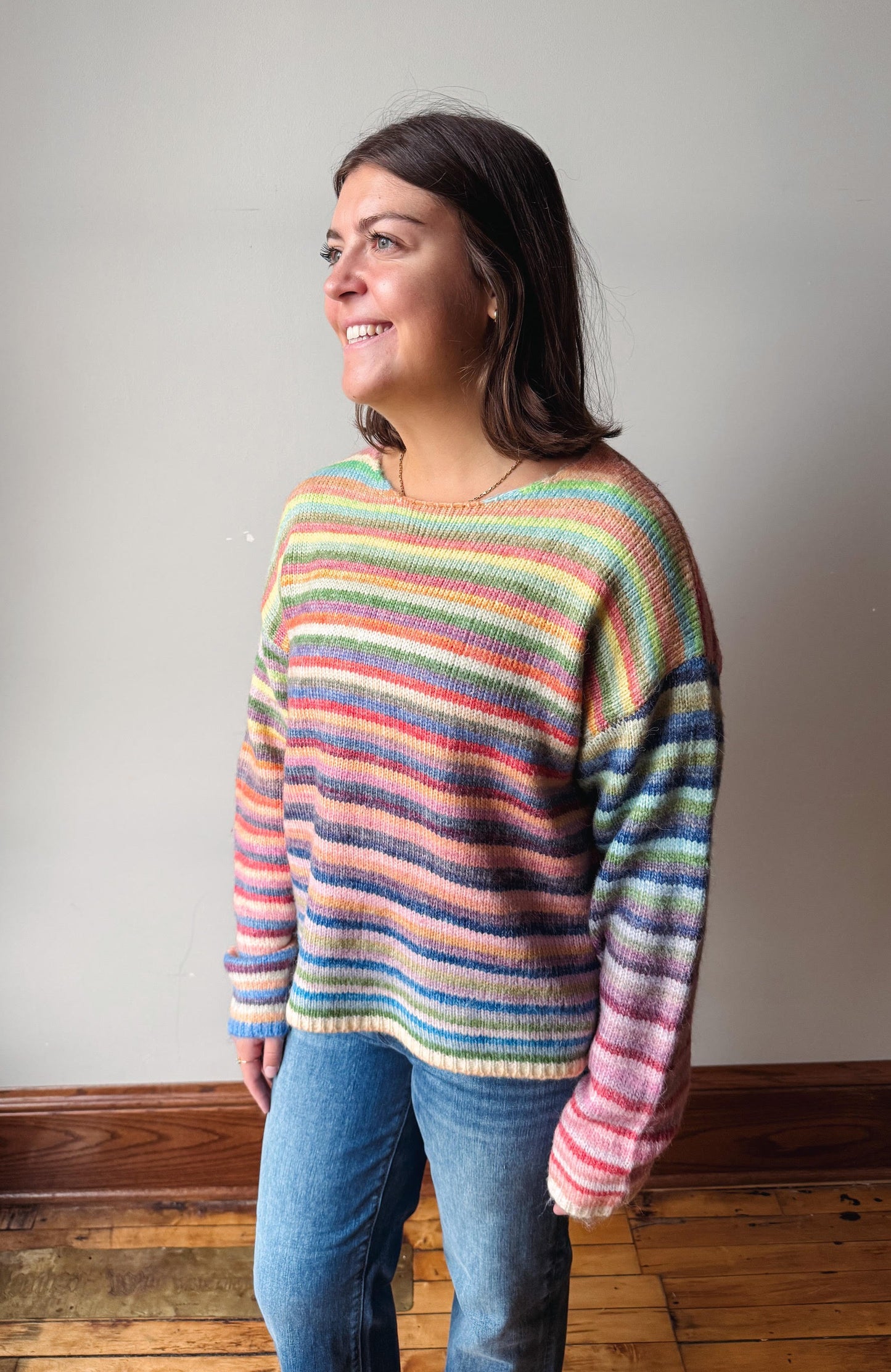A woman wearing a colorful, horizontally striped knit sweater and blue jeans, standing in a room with a light gray wall and wooden floor.