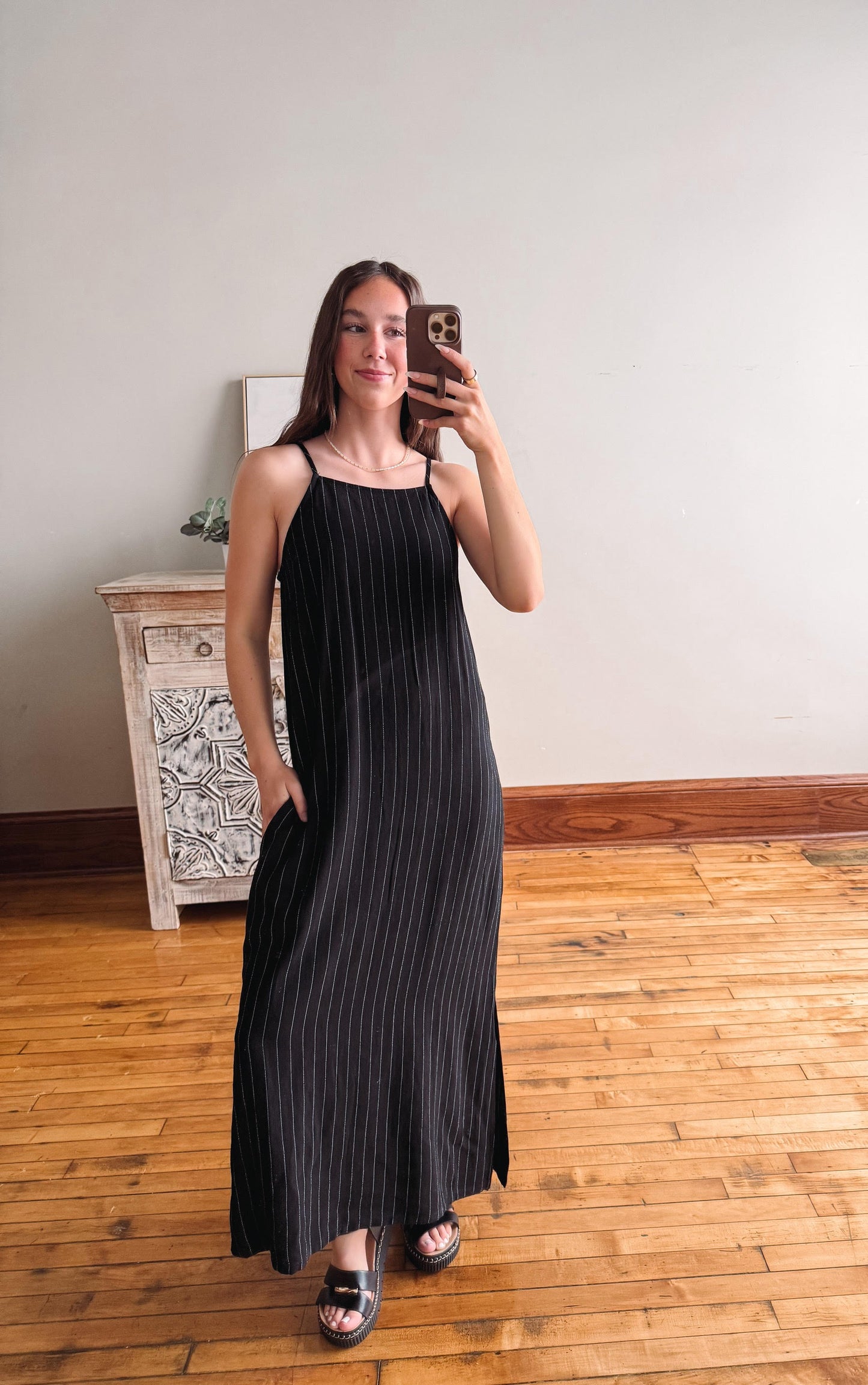 Woman taking a mirror selfie wearing a black sleeveless dress in a room with wooden flooring.