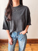 A woman wearing a dark gray knit crewneck sweater with three-quarter sleeves and light blue jeans, standing in front of a plain off-white wall with a wooden floor.