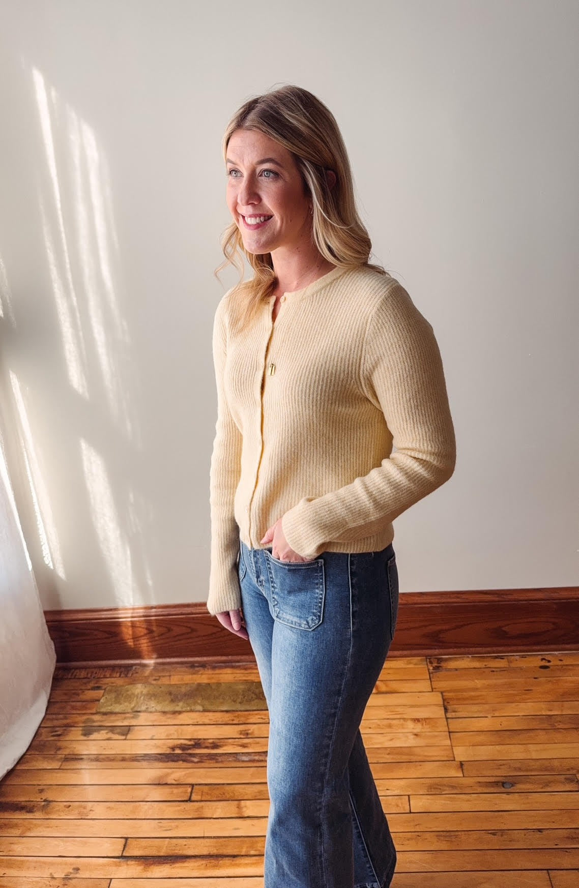 Woman wearing a light yellow cardigan and blue jeans standing in a room with wooden flooring and a white wall.