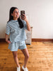 A woman taking a mirror selfie in a room with hardwood floors and a white wall, wearing a matching light blue and white striped short-sleeve t-shirt and shorts set, paired with white sneakers.
