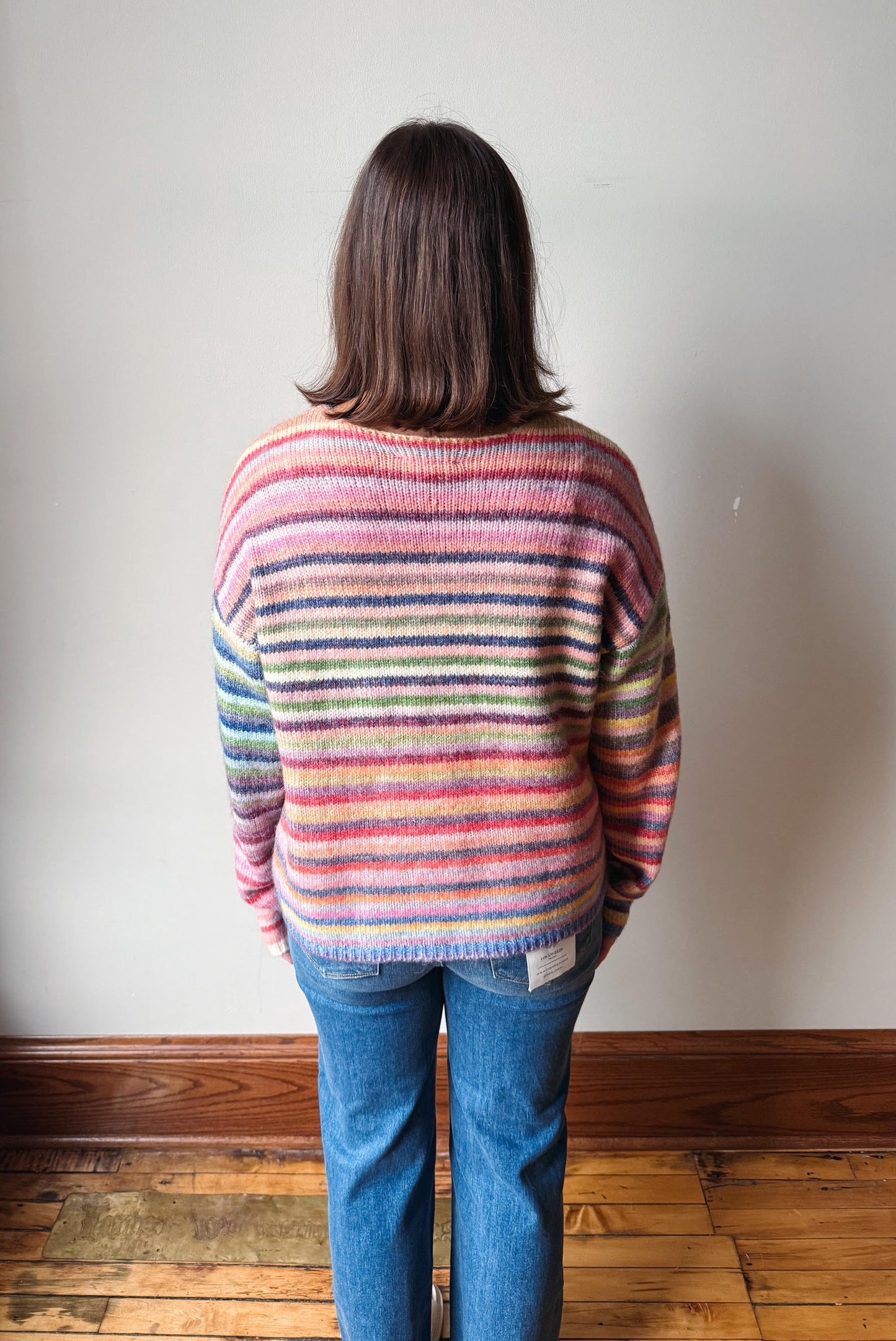 A person viewed from behind wearing a colorful striped knit sweater and blue jeans, standing on a wooden floor against a plain light wall.