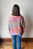A person viewed from behind wearing a colorful striped knit sweater and blue jeans, standing on a wooden floor against a plain light wall.