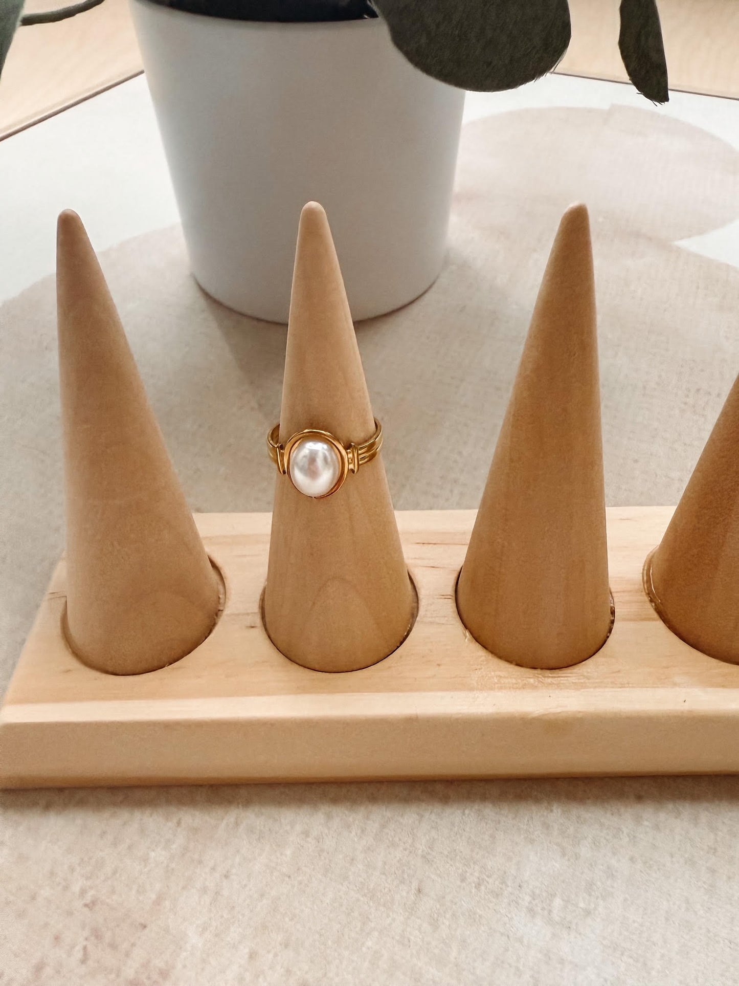 A gold ring with a large white pearl center stone displayed on a light wood cone ring holder, with a white ceramic pot in the background.
