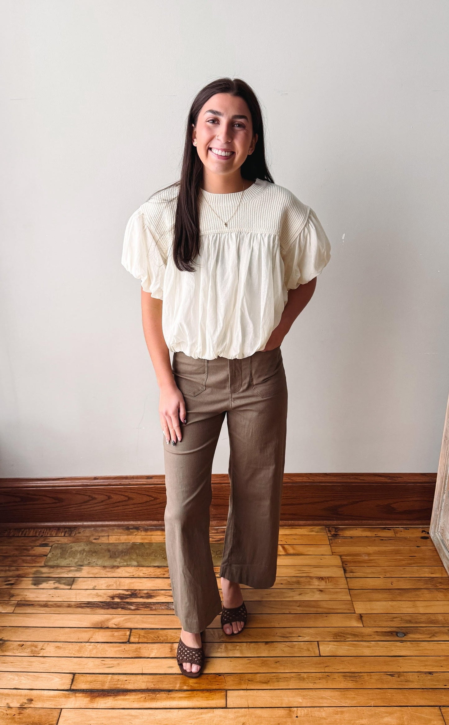 A woman stands in a room with a white wall and wooden floor, wearing a cream puff-sleeve blouse and brown wide-leg pants.
