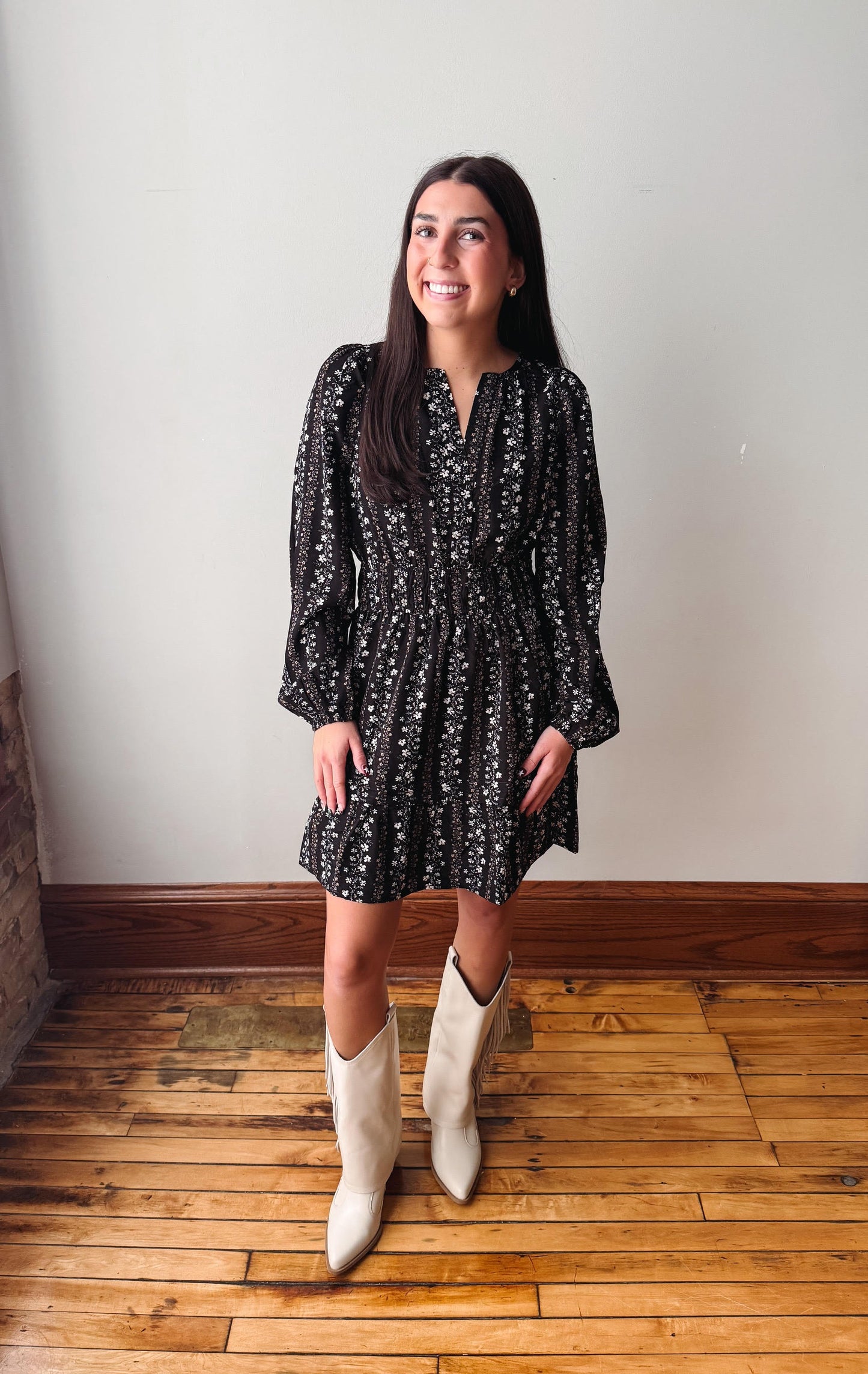 A woman stands indoors wearing a black long-sleeve mini dress with a small white floral pattern and cream-colored cowboy boots, posed against a plain off-white wall on a wooden floor.