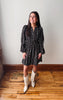 A woman stands indoors wearing a black long-sleeve mini dress with a small white floral pattern and cream-colored cowboy boots, posed against a plain off-white wall on a wooden floor.