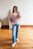 A woman takes a mirror selfie wearing a pastel striped knit sweater, blue flare jeans, and white slide sandals in a room with a light gray wall and wooden floor.
