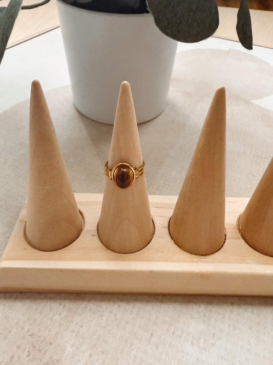 A gold ring with a round brown cabochon stone displayed on a light wood cone ring holder, set on a light beige fabric surface with a white pot in the background.
