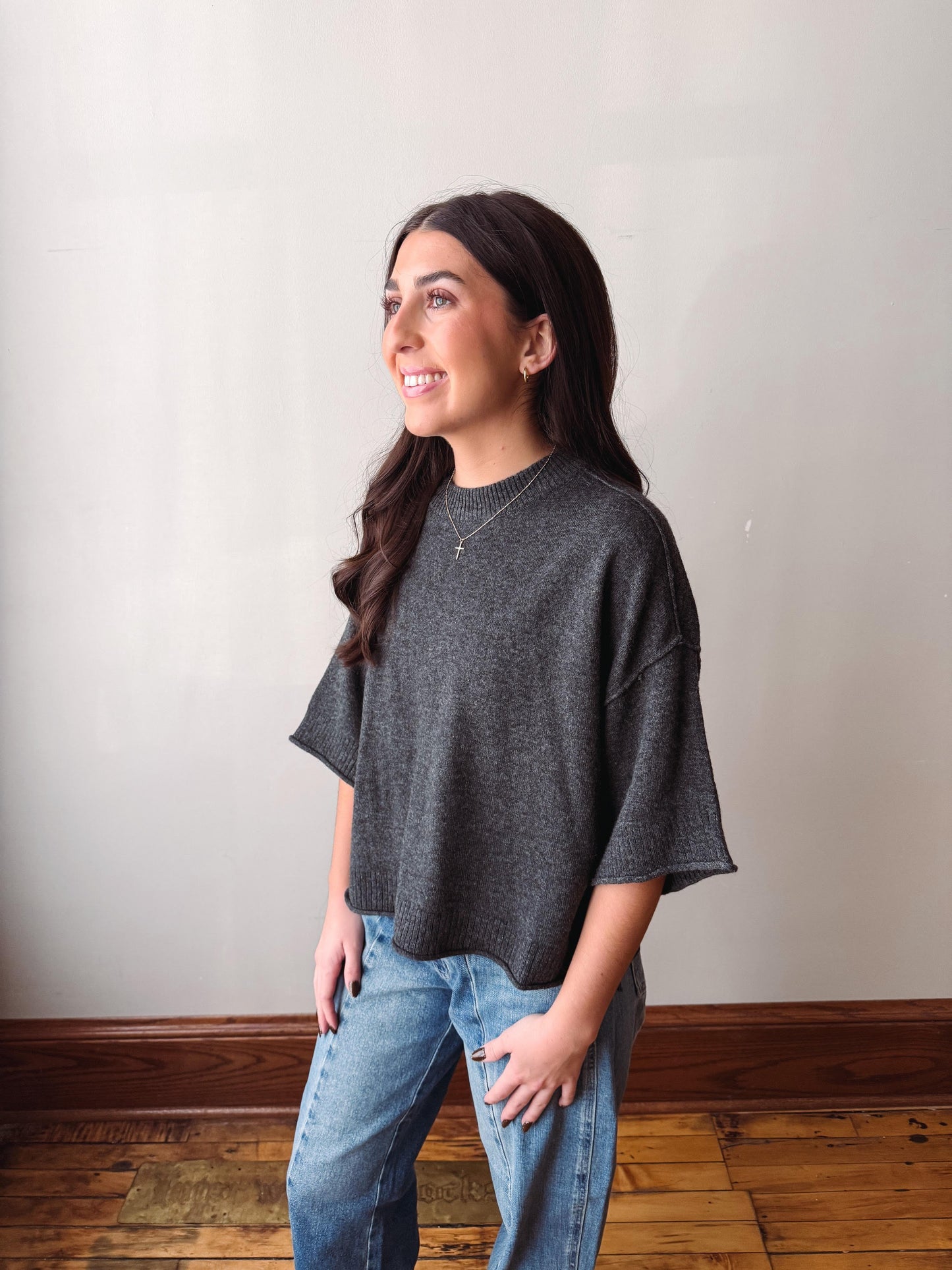 A woman wearing a dark gray heathered short-sleeve knit top and light blue jeans, standing in a room with a beige wall and wooden floor.