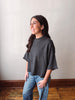 A woman wearing a dark gray heathered short-sleeve knit top and light blue jeans, standing in a room with a beige wall and wooden floor.