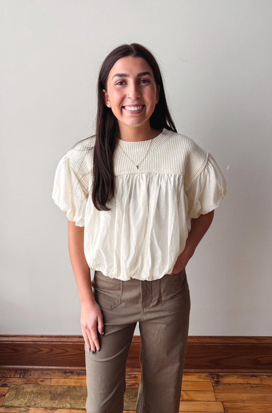 A smiling woman with long dark hair stands indoors wearing a cream-colored short-sleeve blouse with a gathered, ruffled neckline and taupe-brown pants against a plain light gray wall.