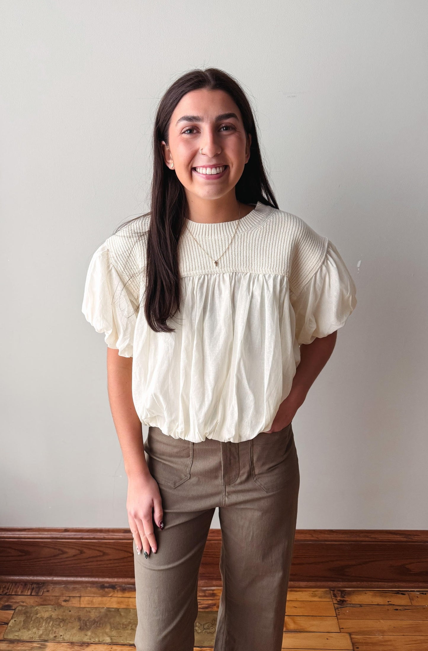 A smiling woman with long dark hair stands indoors wearing a cream-colored short-sleeve blouse with a gathered, ruffled neckline and taupe-brown pants against a plain light gray wall.