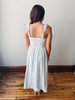 Woman wearing a white dress with thin straps and ribbon details, standing on a wooden floor against a plain wall.