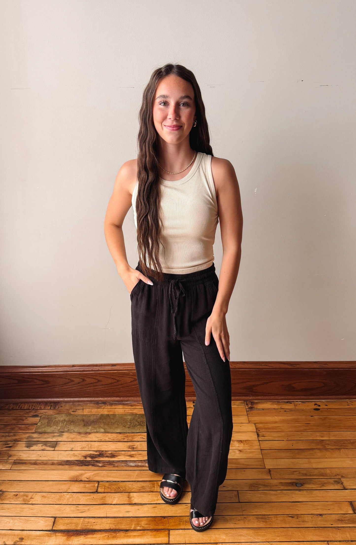 Woman wearing a beige sleeveless top and black pants standing on a wooden floor with a white wall background.