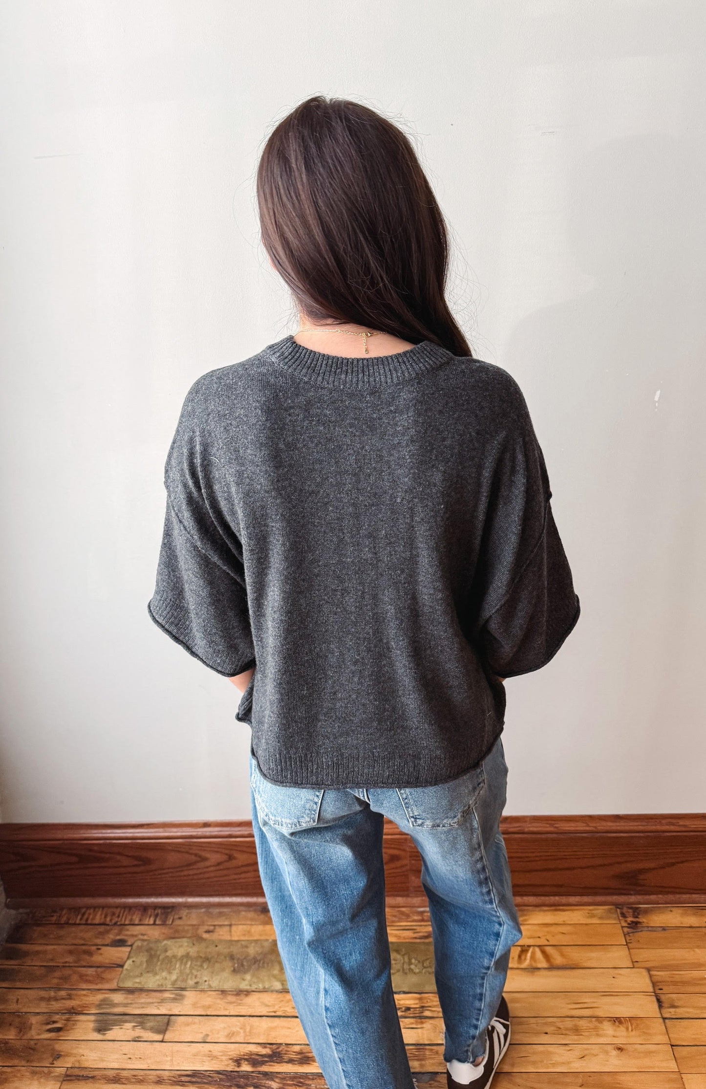 A woman viewed from behind wearing a dark gray knit sweater and blue jeans, standing on a wooden floor against a plain off-white wall.