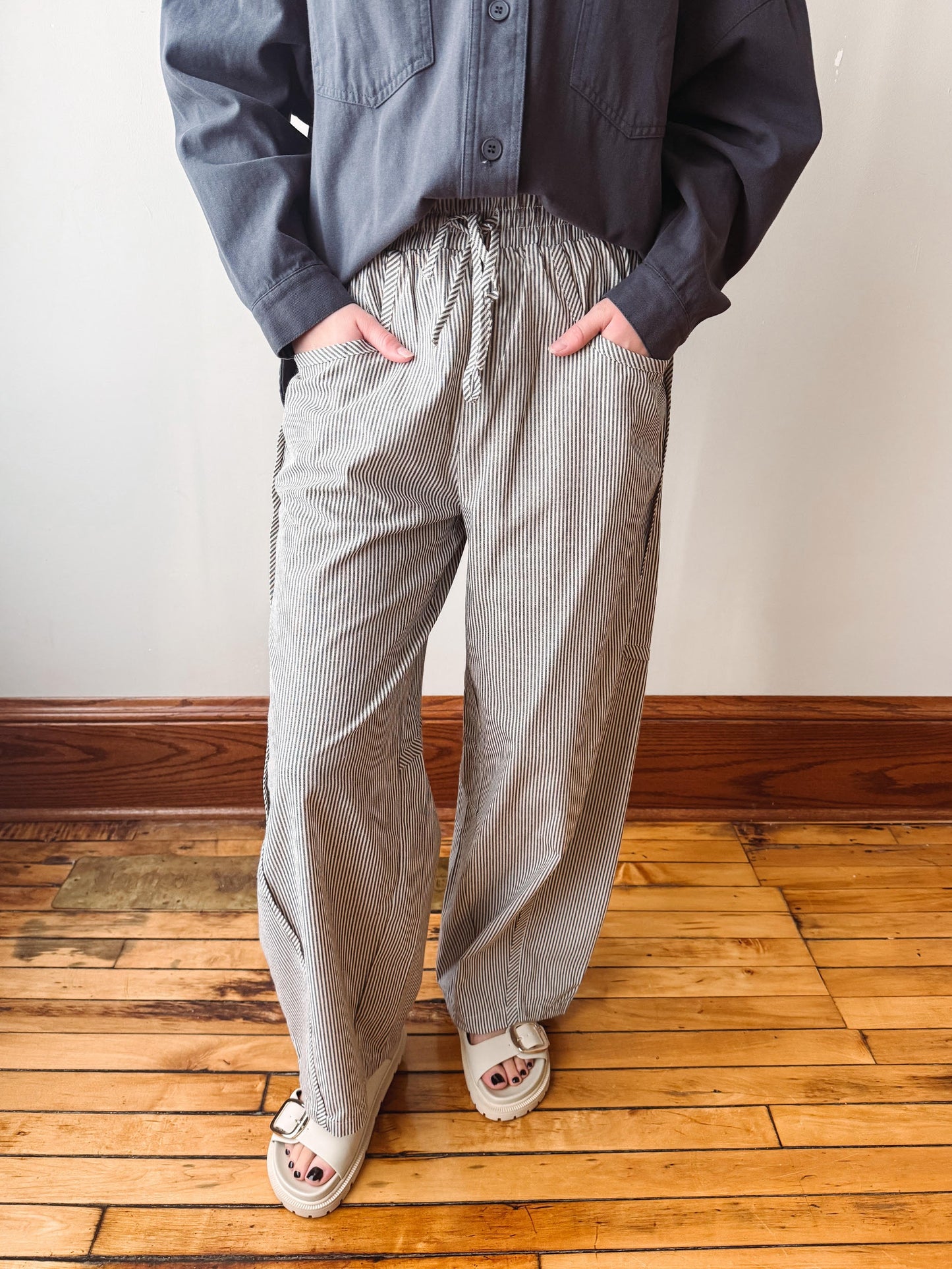 A person wearing loose, wide-leg pinstriped trousers in a gray and white pattern, paired with a dark gray button-up shirt and white slide sandals, standing on a wooden floor against a plain wall.