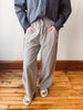 A person wearing loose, wide-leg pinstriped trousers in a gray and white pattern, paired with a dark gray button-up shirt and white slide sandals, standing on a wooden floor against a plain wall.