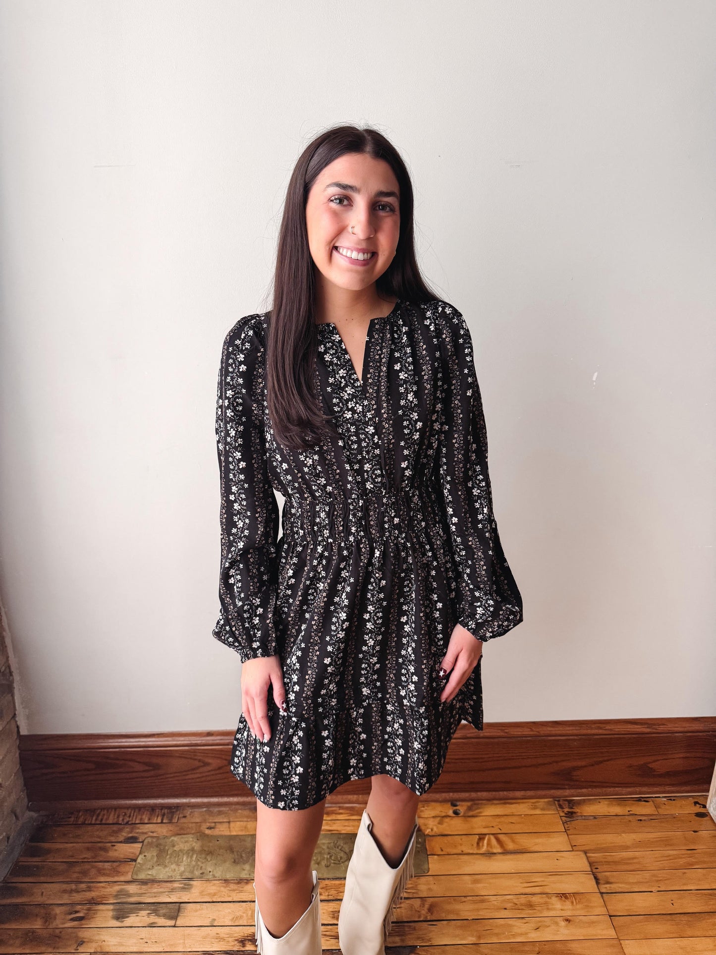 A woman wearing a black long-sleeve mini dress with a white floral pattern, standing in front of a plain off-white wall on a wooden floor.