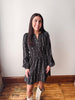 A woman wearing a black long-sleeve mini dress with a white floral pattern, standing in front of a plain off-white wall on a wooden floor.