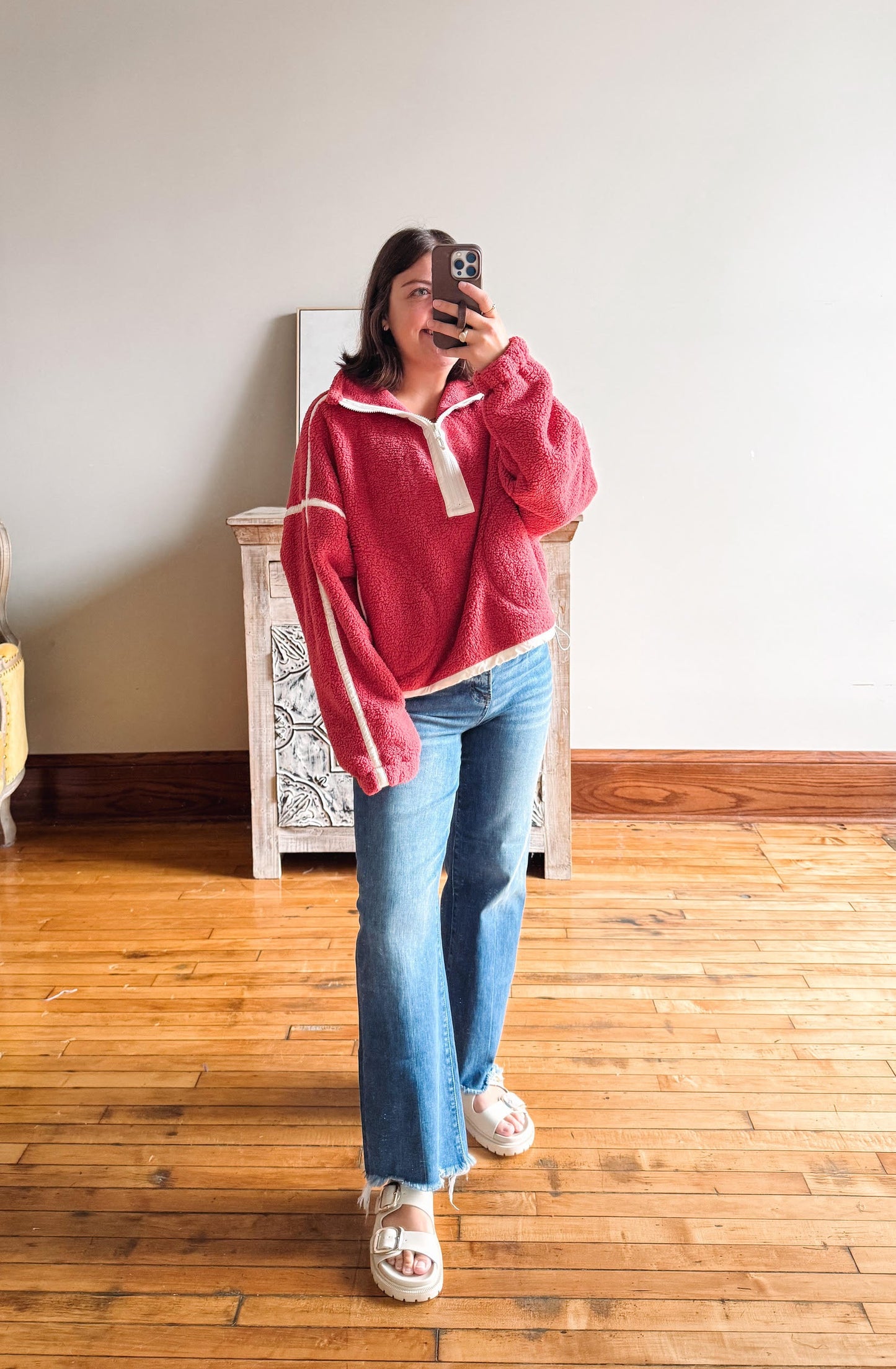 A woman takes a mirror selfie wearing a fuzzy pink quarter-zip fleece pullover with cream trim, blue flare jeans, and white slide sandals in a room with a wooden floor and light-colored wall.