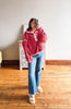 A woman takes a mirror selfie wearing a fuzzy pink quarter-zip fleece pullover with cream trim, blue flare jeans, and white slide sandals in a room with a wooden floor and light-colored wall.