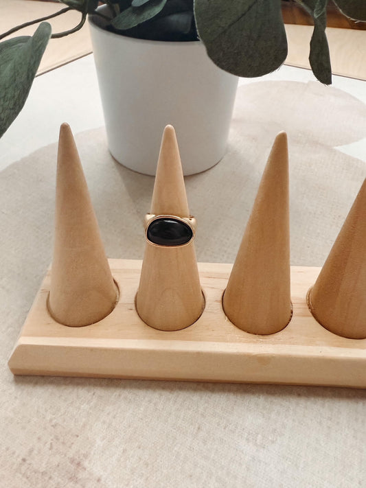 A gold ring with a round black cabochon stone displayed on a light wood cone ring holder, set on a beige fabric surface with a white planter in the background.