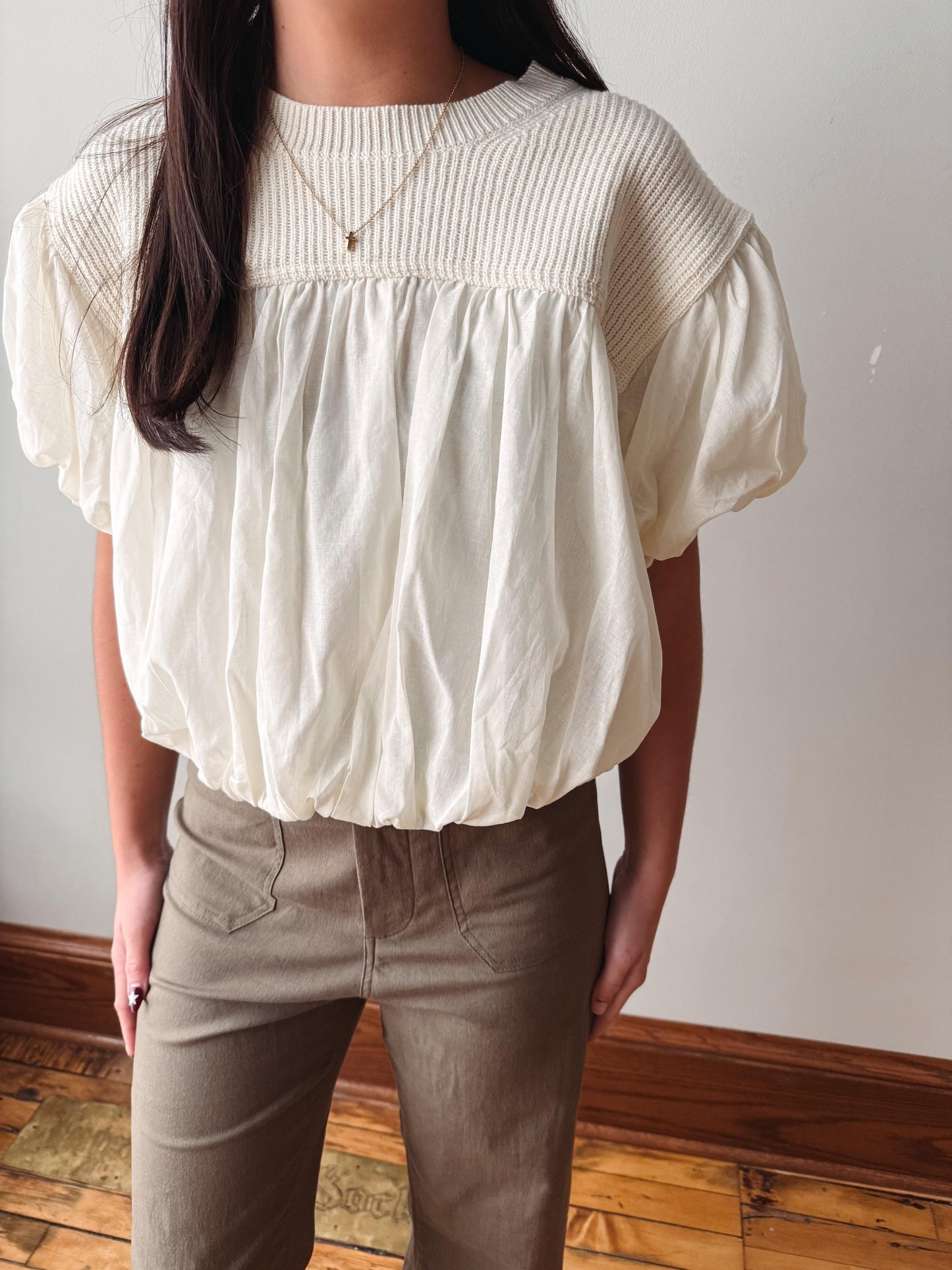 A woman wearing a cream-colored short-sleeve blouse with a smocked yoke and gathered puff sleeves, paired with brown pants, standing indoors against a plain wall.