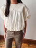 A woman wearing a cream-colored short-sleeve blouse with a smocked yoke and gathered puff sleeves, paired with brown pants, standing indoors against a plain wall.