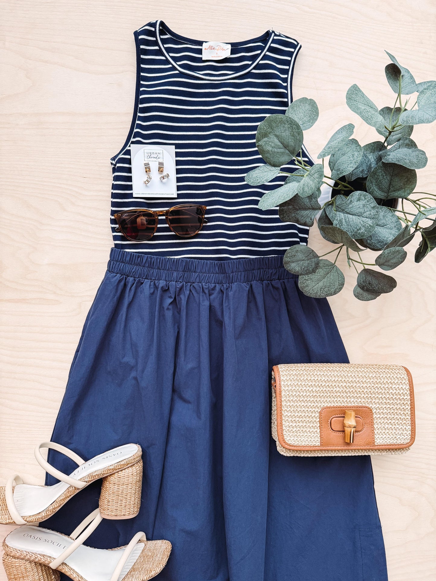 Navy blue dress with striped top, sunglasses, sandals, and a woven handbag on a light background.