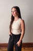 Woman wearing a beige sleeveless top and black pants against a plain wall.