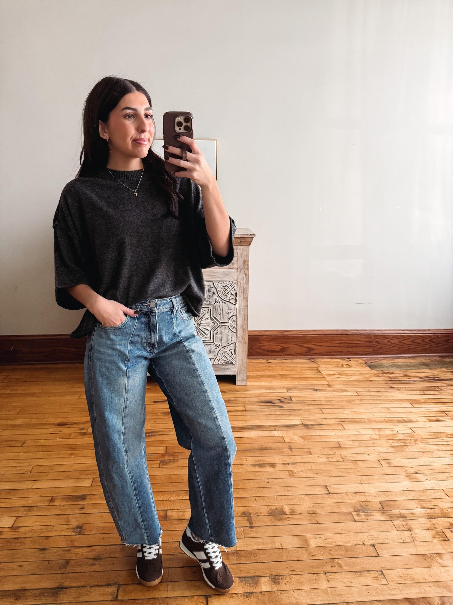 A woman taking a mirror selfie in a room with light-colored walls and a wooden floor, wearing a dark gray t-shirt, wide-leg blue jeans, and brown sneakers.
