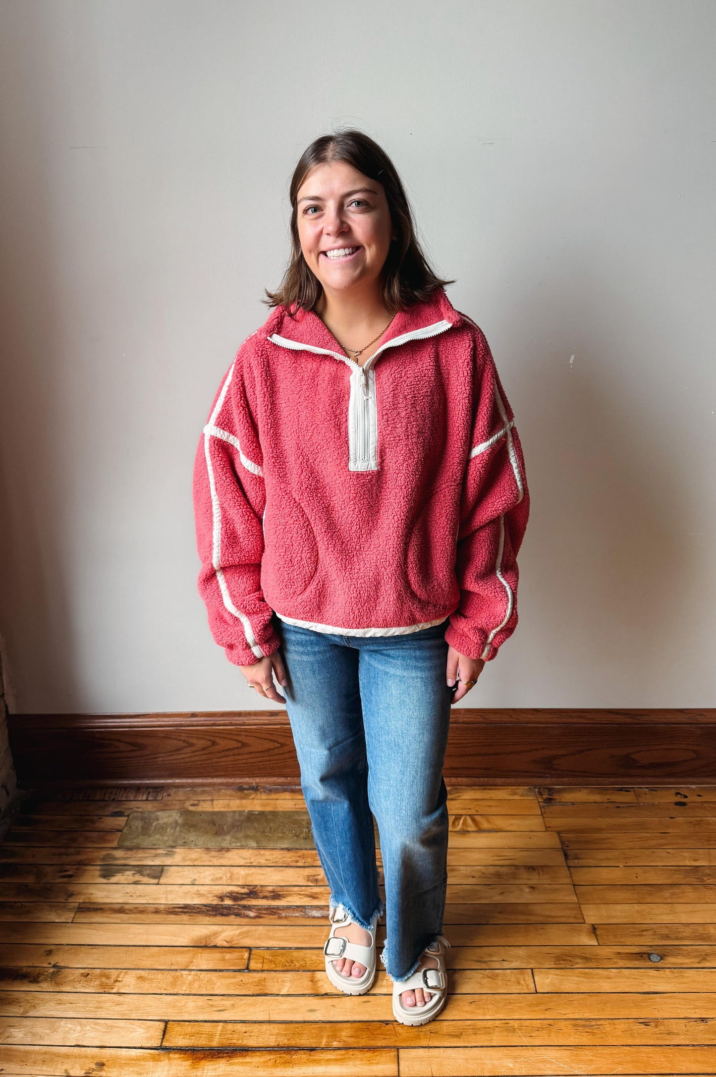 A woman wearing a pink fleece pullover with white trim, blue jeans, and white slide sandals, standing on a wooden floor against a plain wall.