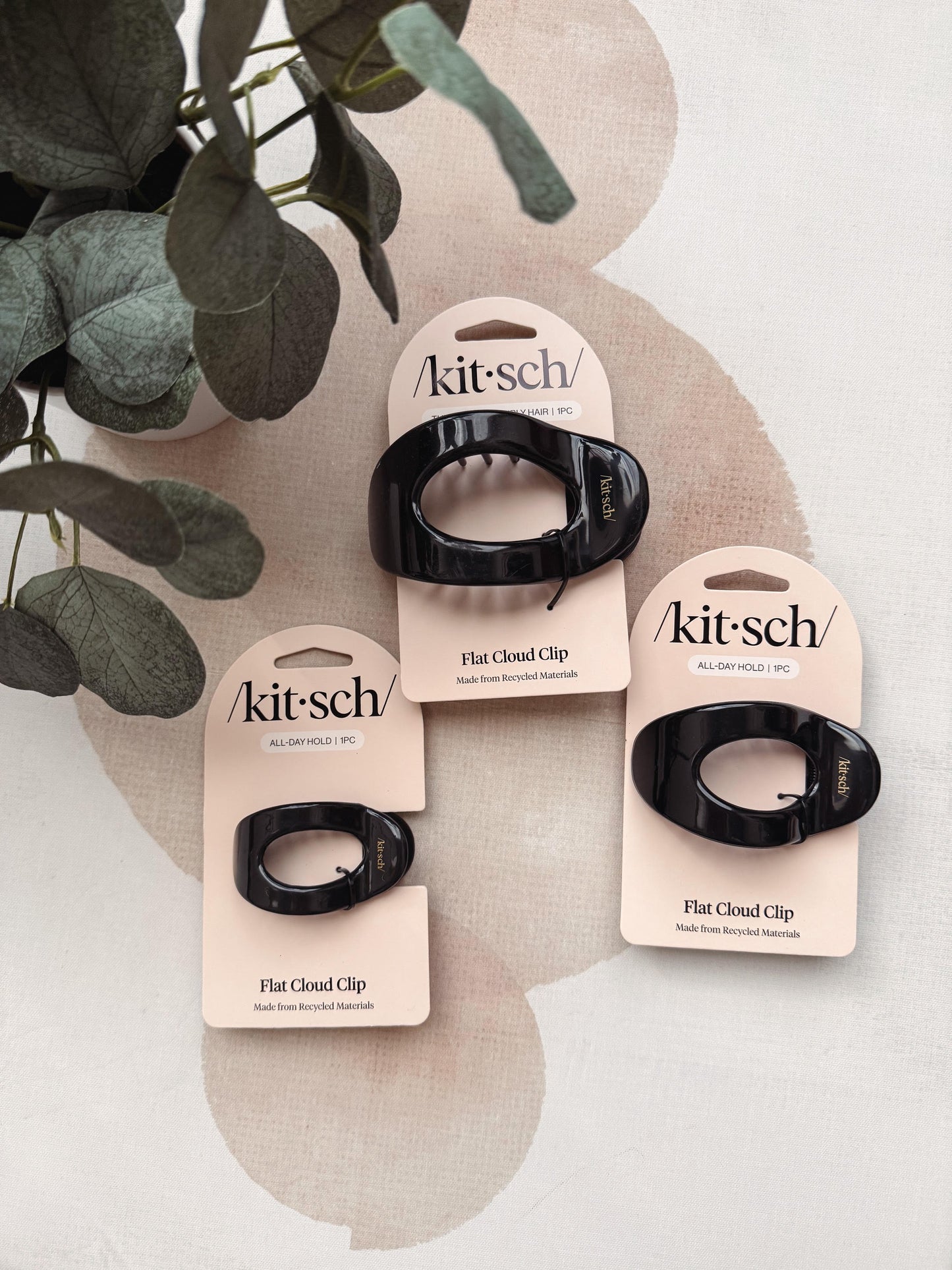 Three black flat cloud hair clips on pink retail cards, styled with green leaves on a light beige surface.