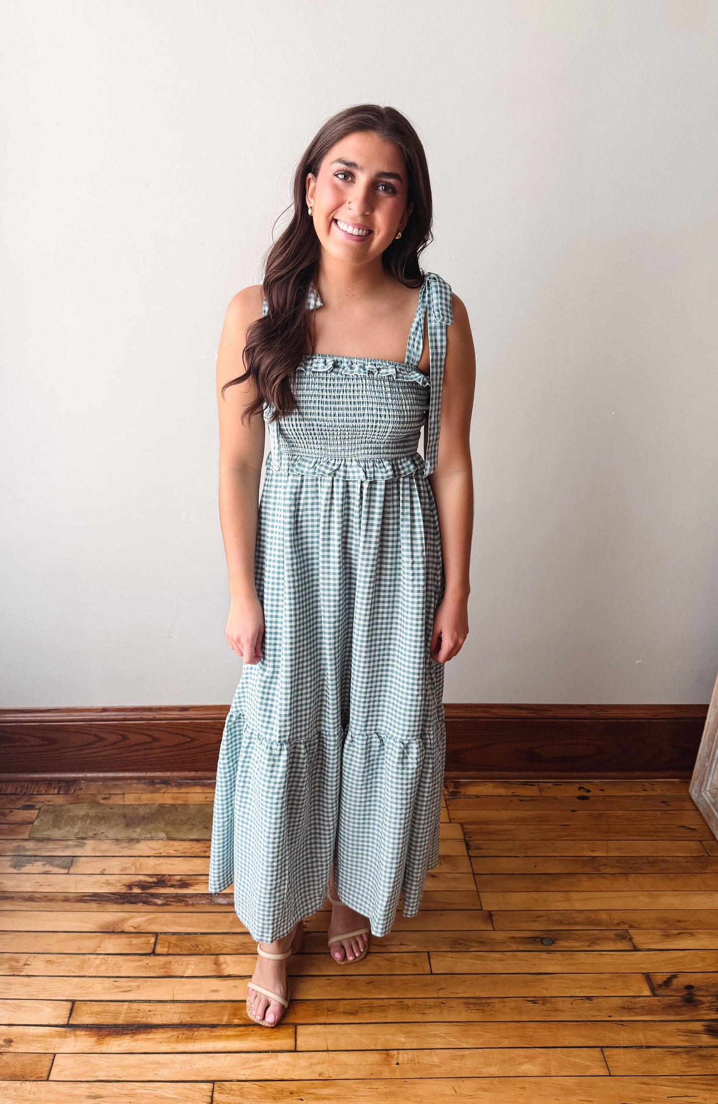Woman wearing a green checkered dress standing on a wooden floor with a white wall background