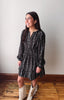 A woman modeling a black long-sleeve mini dress with a white floral pattern, paired with beige fringe boots, standing on a wooden floor against a plain light gray wall.