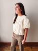 A woman modeling a cream-colored short-sleeve blouse with textured vertical pleats and puff sleeves, paired with khaki pants, standing against a neutral wall.