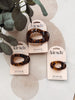 Three Kit'sch flat cloud hair clips in a tortoiseshell pattern, each on its original cream-colored card packaging, styled with green leaves on a light beige textured background.