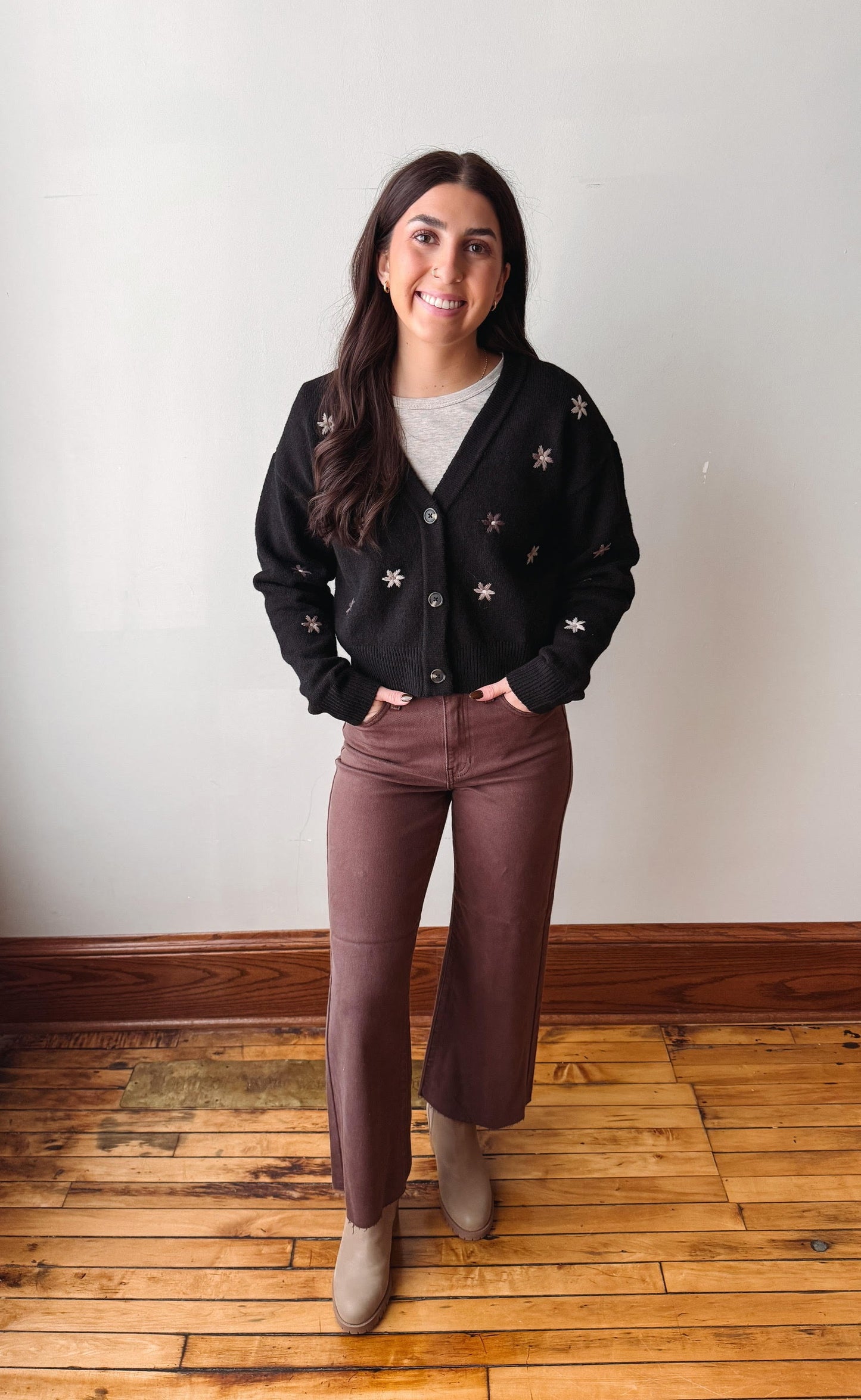 A woman stands in a room with a white wall and wooden floor, wearing a black button-up cardigan with white embroidered star details, brown wide-leg pants, and beige ankle boots.