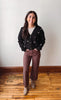 A woman stands in a room with a white wall and wooden floor, wearing a black button-up cardigan with white embroidered star details, brown wide-leg pants, and beige ankle boots.