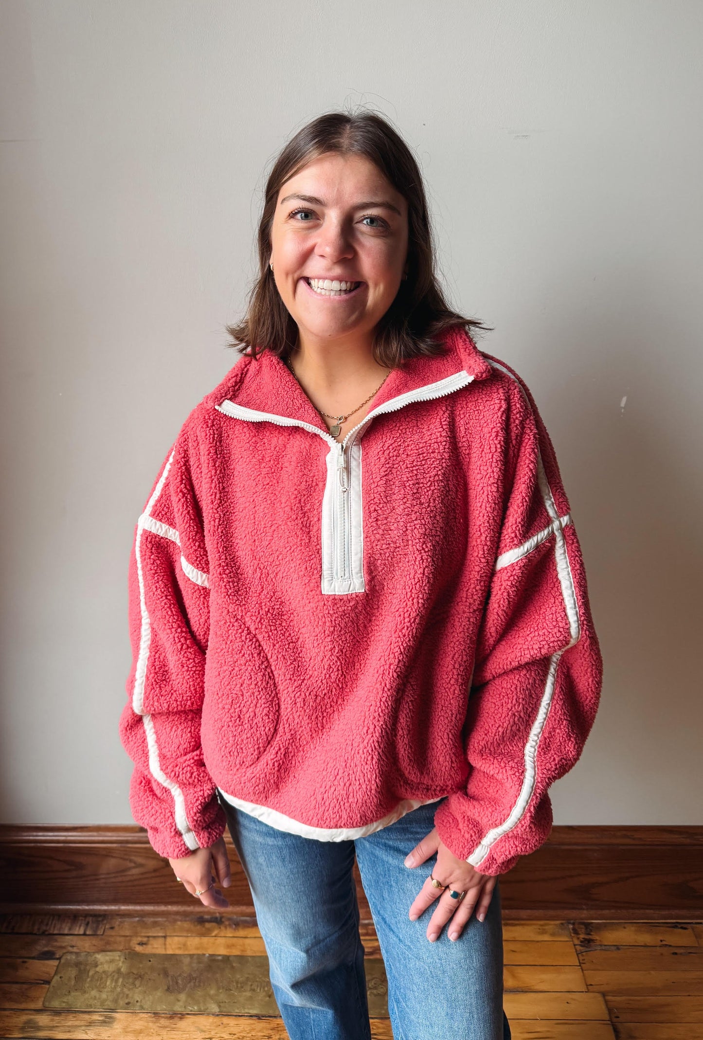 A smiling woman wearing a pink fleece quarter-zip pullover with white contrast trim and blue jeans, standing against a plain light gray wall with a wooden floor visible.