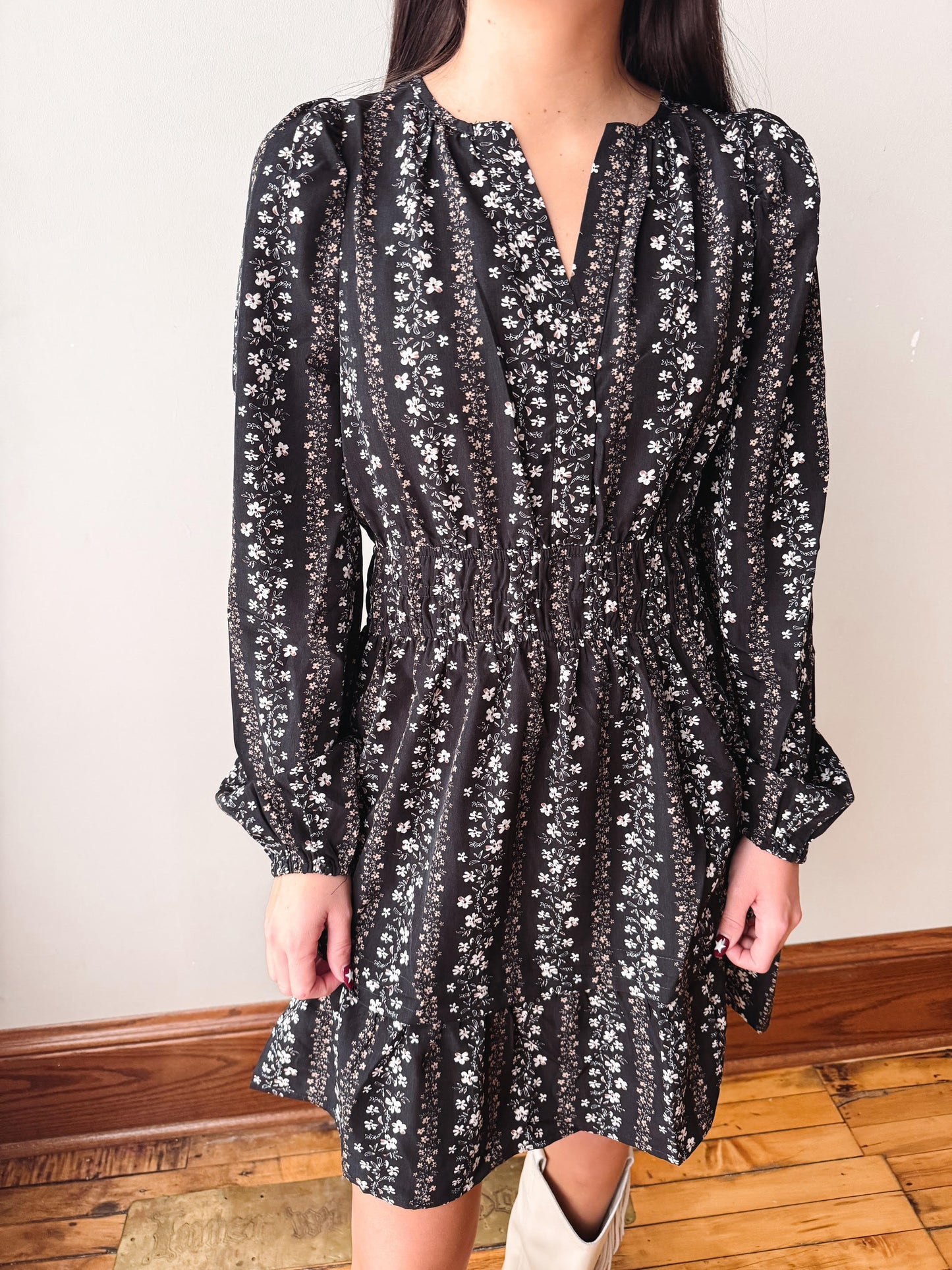 A woman wearing a black long-sleeve mini dress with a white floral print, standing in an indoor setting with a light-colored wall and wooden floor visible.